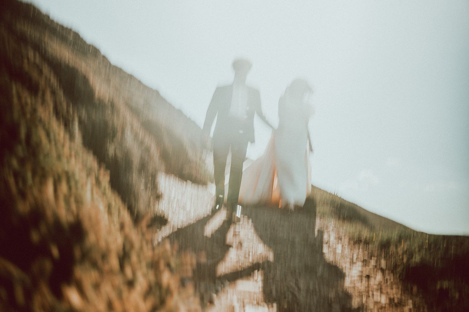 A couple in wedding attire holds hands while walking along a sunlit sandy dune in a dreamy, soft-focus photograph.