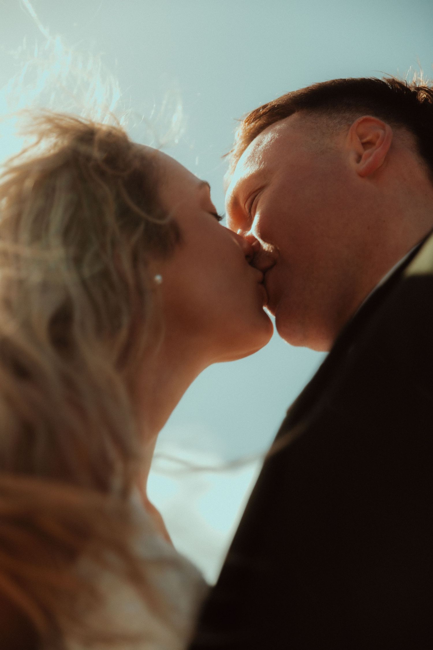 Close-up of a couple kissing outdoors under a bright blue sky, with the woman's wind-blown blonde hair glowing in warm sunlight.