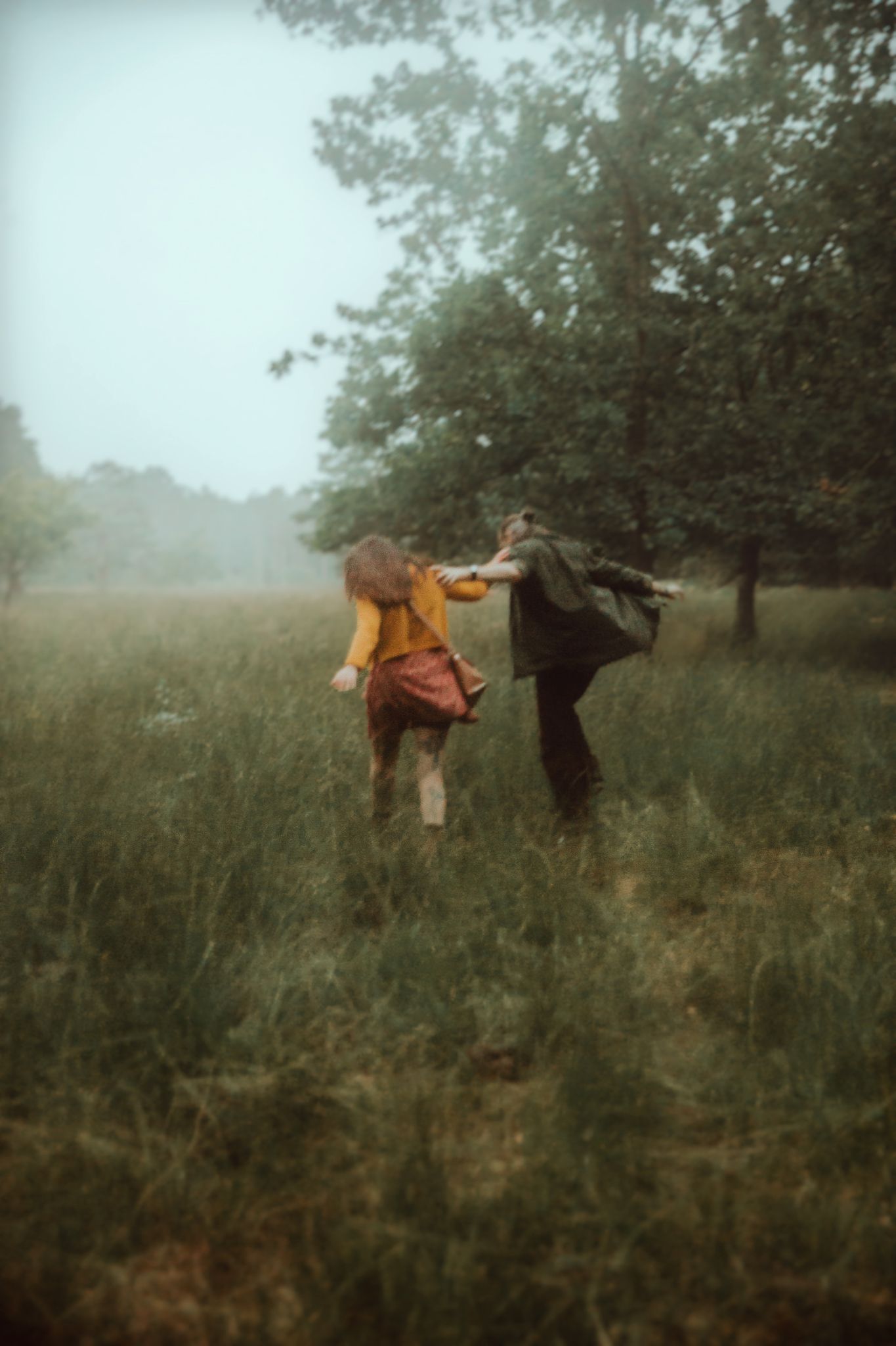 Two people in dark clothing walk through a misty field at dusk with an ethereal atmospheric quality.