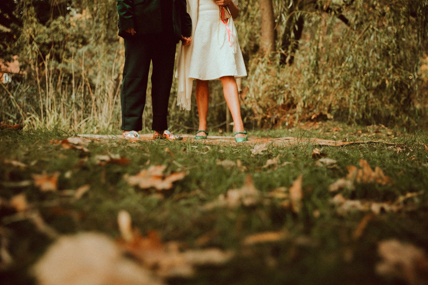 Couple standing together on grass with autumn leaves, wearing casual attire, shot from waist down in a romantic outdoor setting.