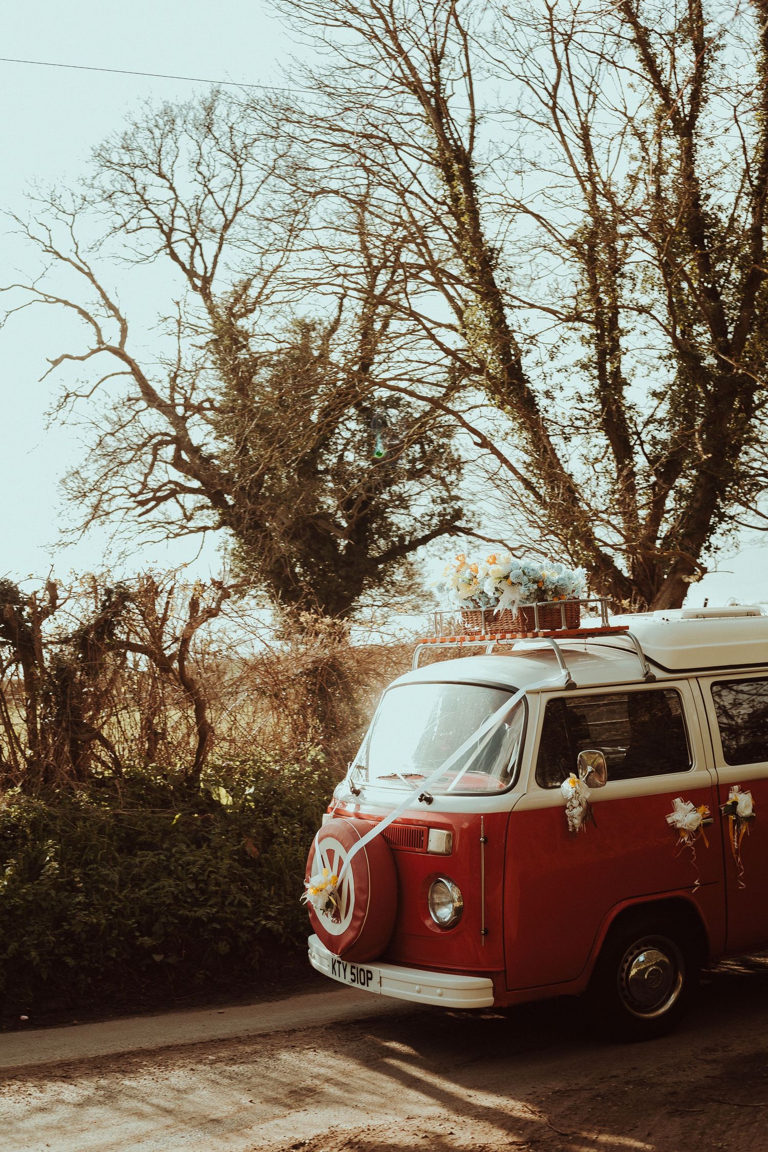 A vintage red Volkswagen camper van decorated with flowers and ribbons parked on a road surrounded by bare trees.