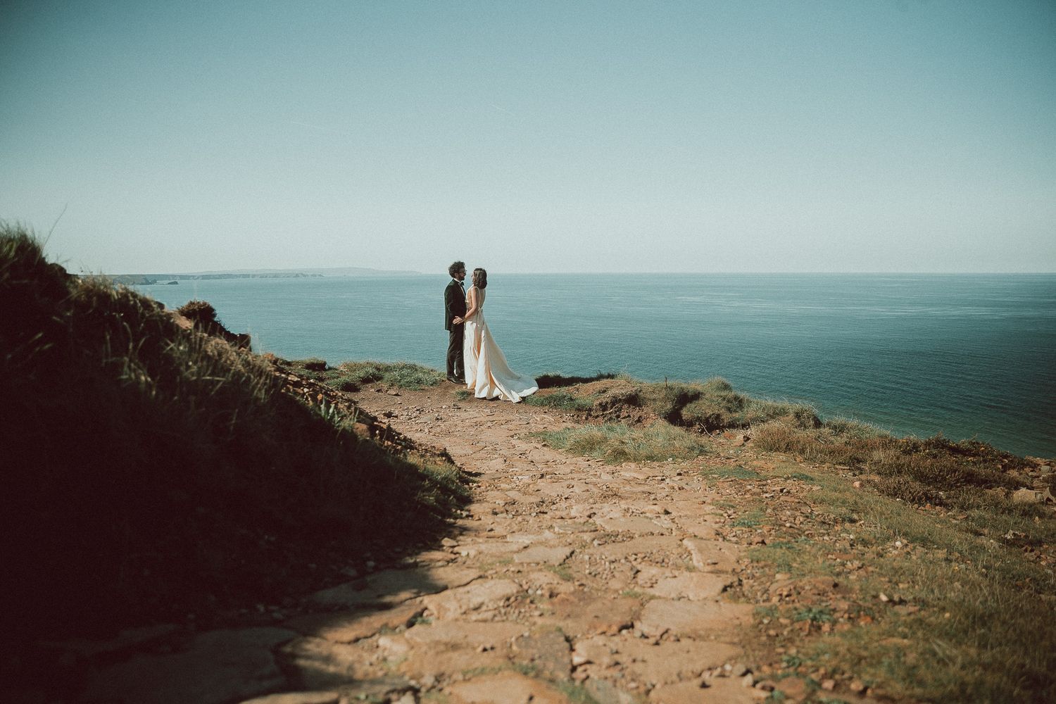 Two silhouettes stand on a coastal cliff path overlooking a serene blue ocean on a misty day.
