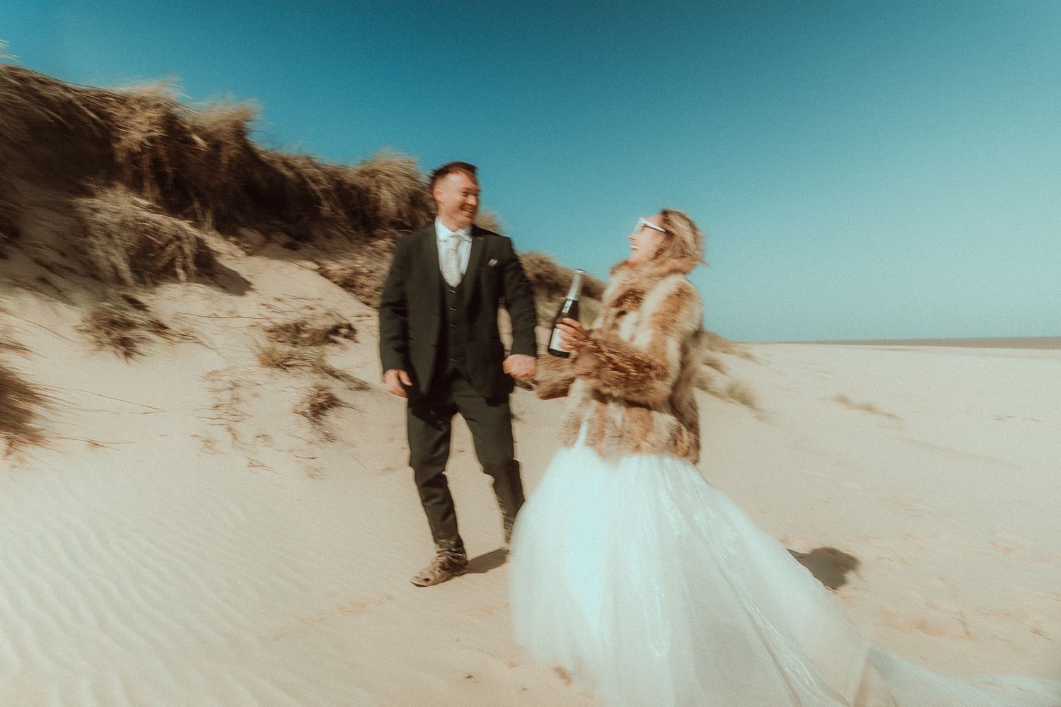 Bride and groom walking hand in hand on sandy beach dunes, bride in white gown with fur stole, sunny blue sky.