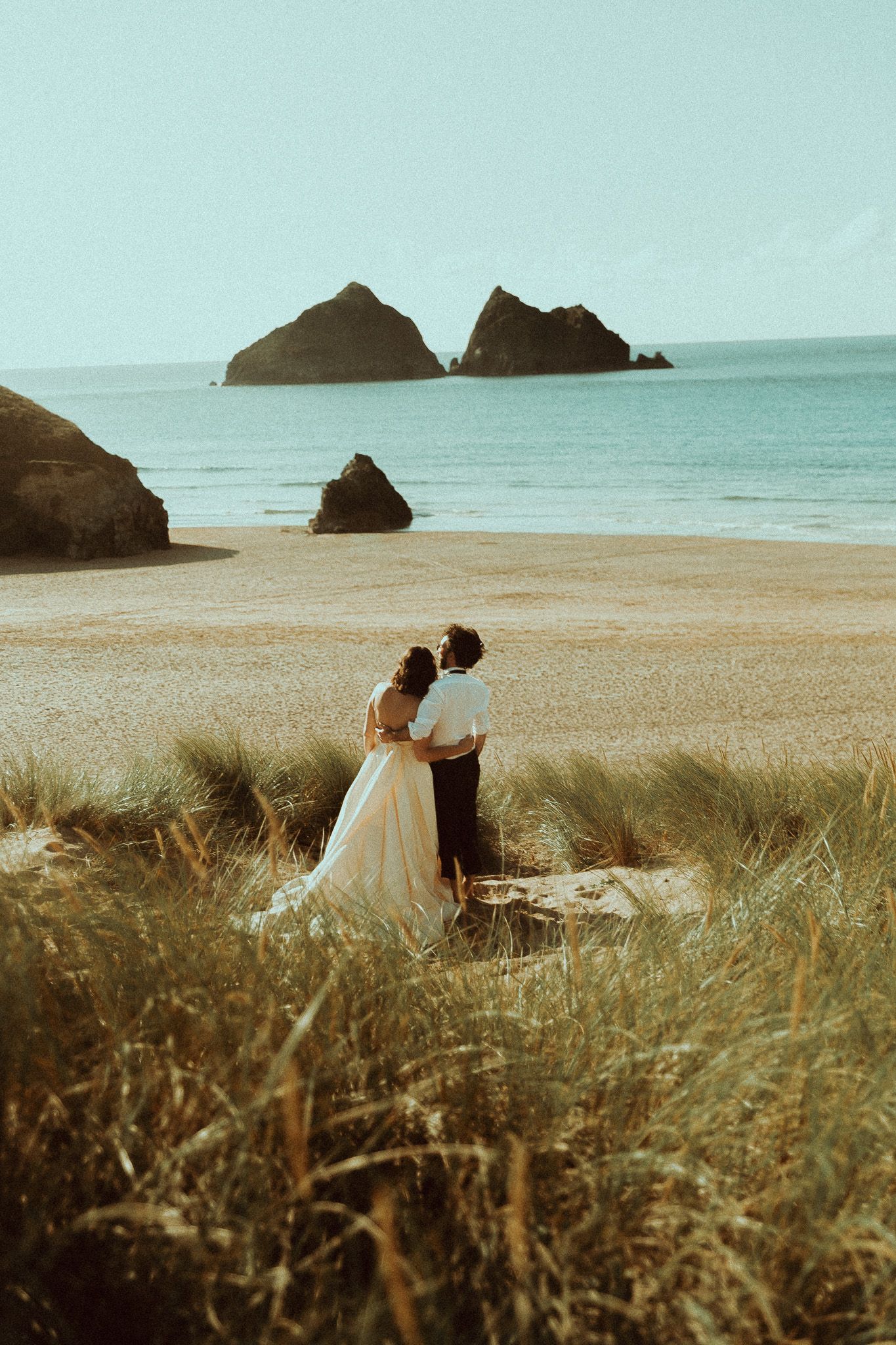 A couple embraces on a sandy beach with dramatic coastal rock formations and turquoise waters in the background.