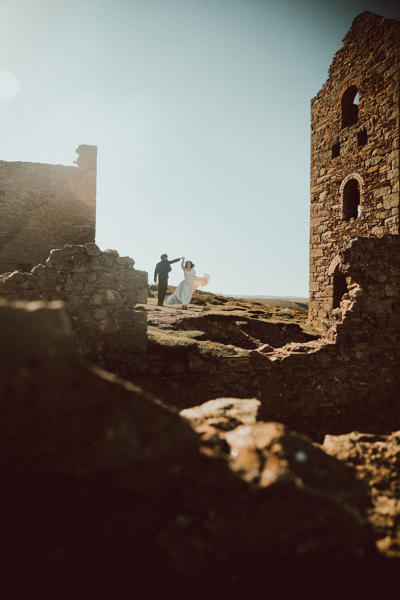 Bride in a flowing white dress dances with her partner among ancient stone ruins on a sunlit hilltop during a romantic elopement.