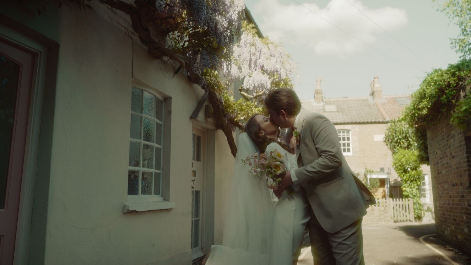 A couple shares a romantic kiss outside a charming cottage adorned with wisteria, bride holding a bouquet.