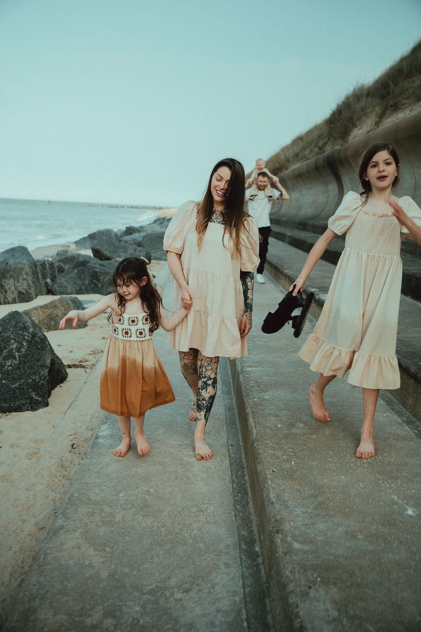Children in light dresses play and explore barefoot along a rocky coastal path on a misty day.