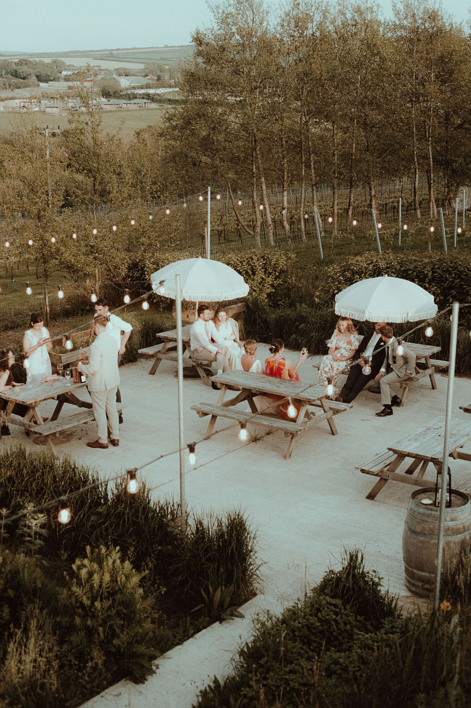 Guests socialising at outdoor picnic tables with white umbrellas and string lights at a countryside wedding venue.