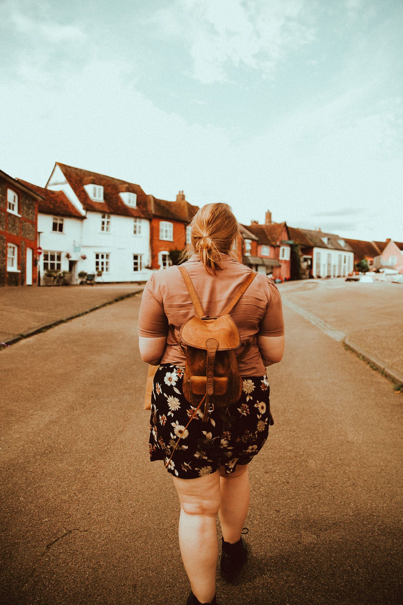 A woman with a brown leather backpack walks down a quiet street in Lavenham, Suffolk, lined with traditional red brick English buildings.