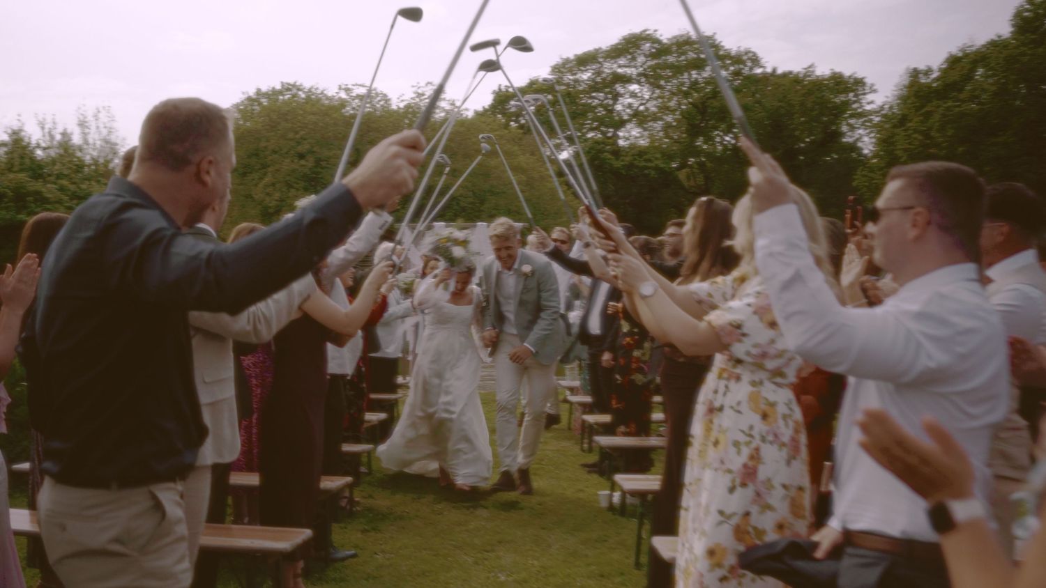 Newlyweds walk through a golf club arch formed by guests at an outdoor wedding ceremony on a green lawn.