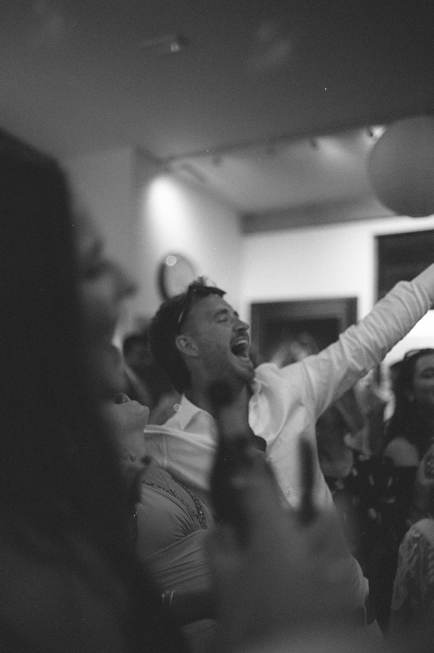 Black and white photo of a man singing and dancing joyfully at a lively indoor party or wedding reception.