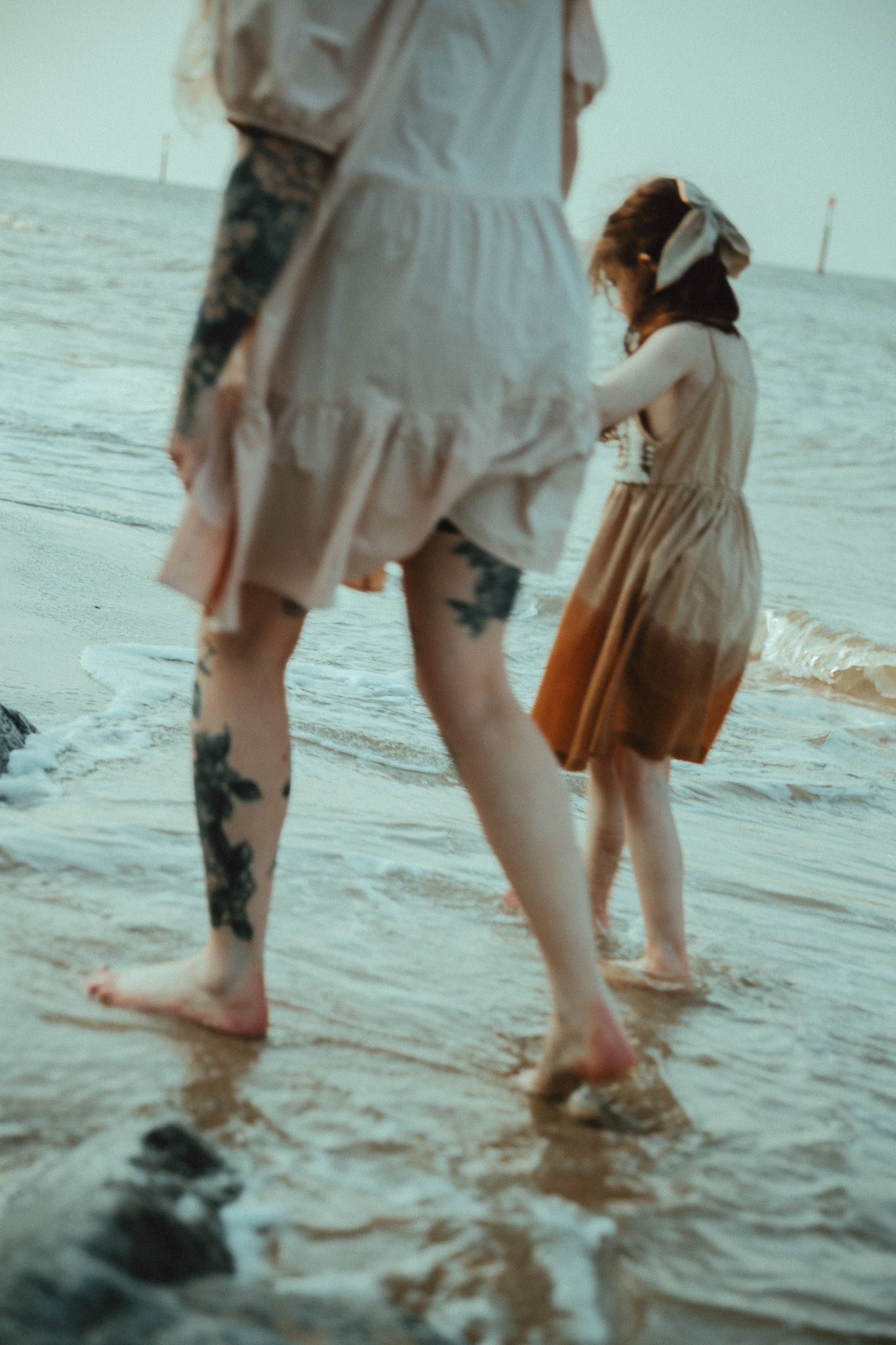 Two people wearing flowy dresses wade in shallow ocean water at sunset, creating a dreamy vintage aesthetic.