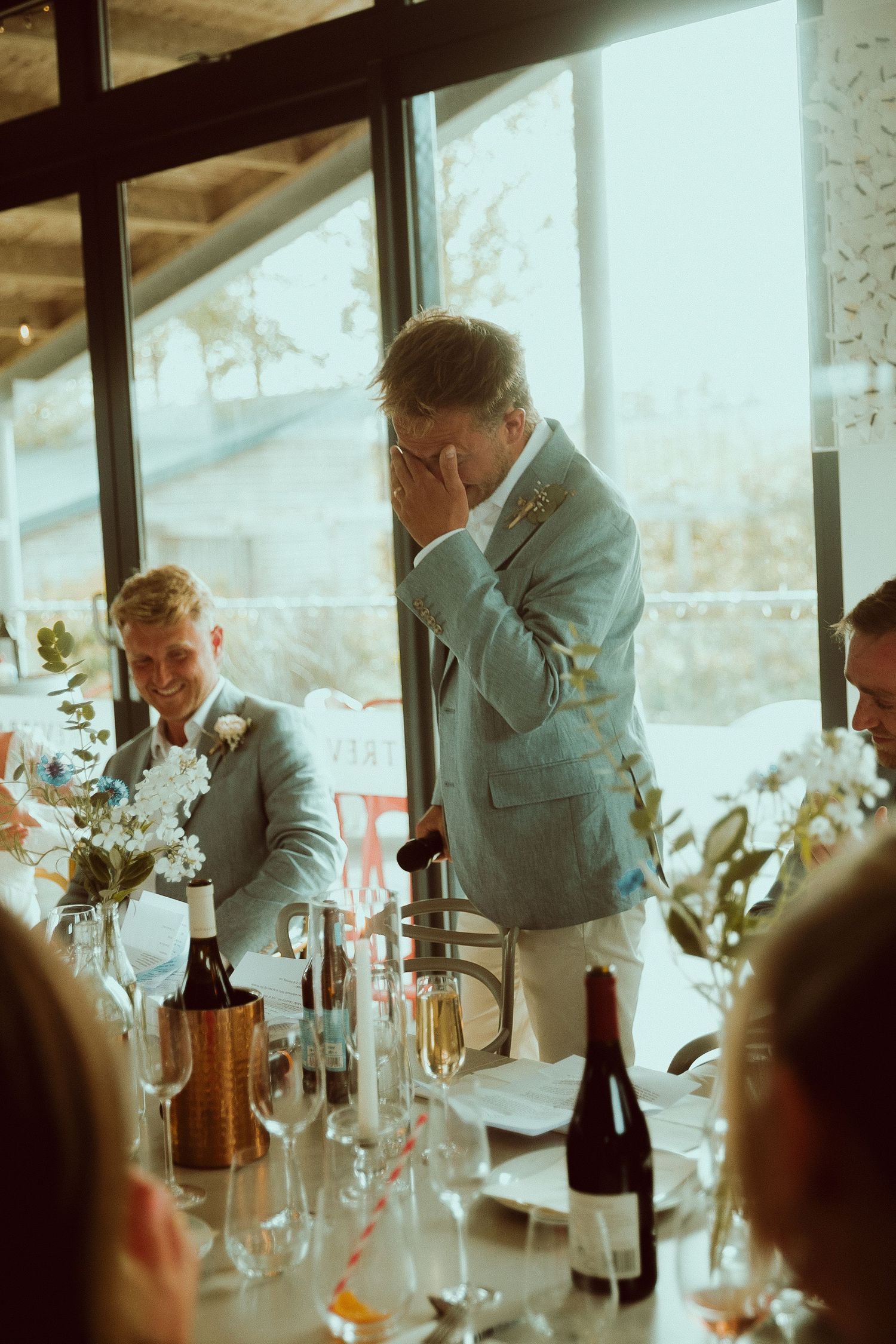 Emotional best man covering his face during wedding reception speech at elegant venue with floral arrangements.