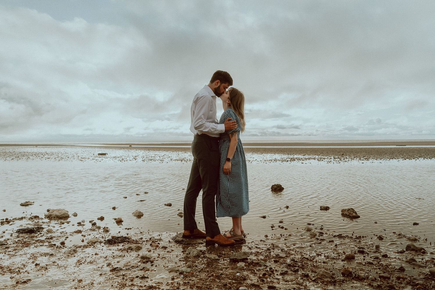 A couple shares a romantic embrace on a rocky beach under dramatic cloudy skies near shallow tidal waters.