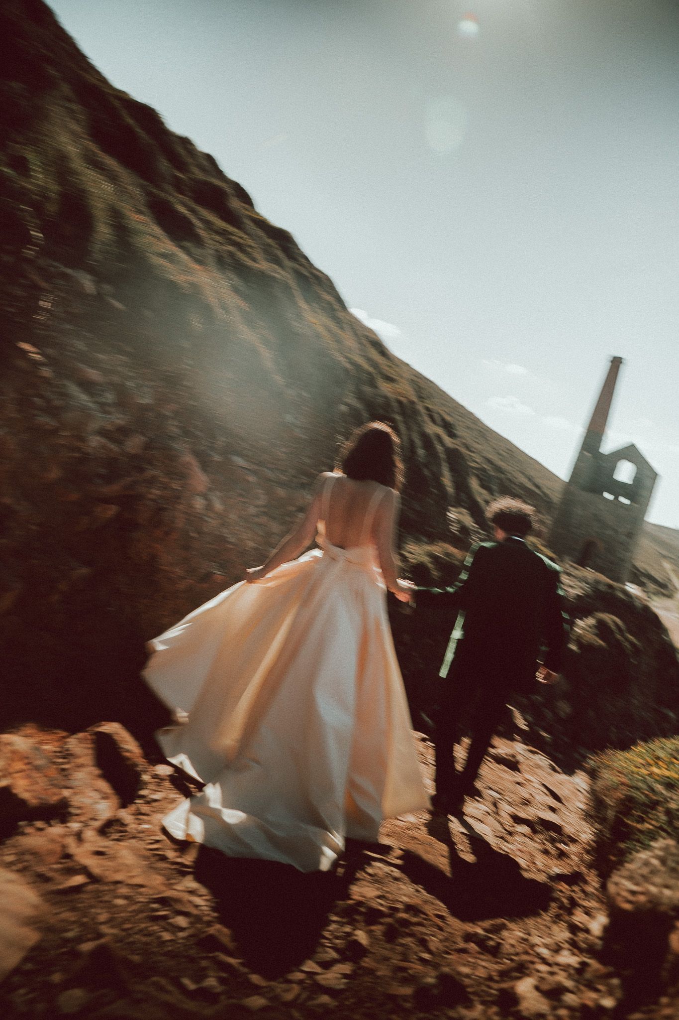 Bride and groom walking hand in hand over rocky terrain near ruins in Cornwall, wedding dress flowing in sunlight.