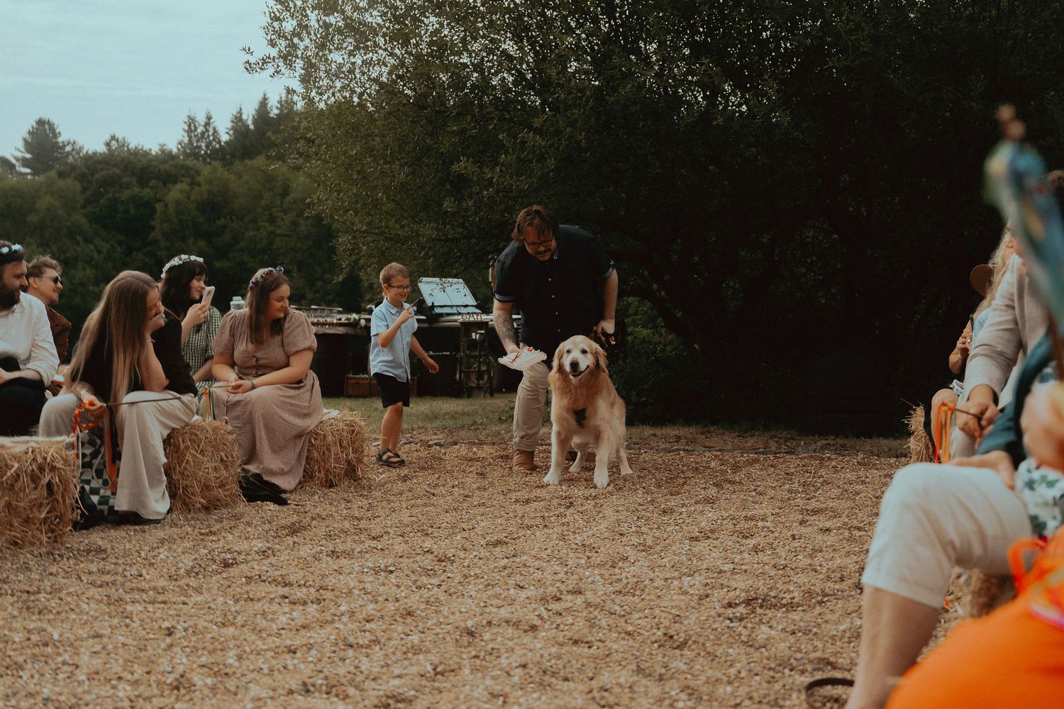 A golden retriever walks with a man at an outdoor gathering, guests seated on hay bales in a rustic field setting.
