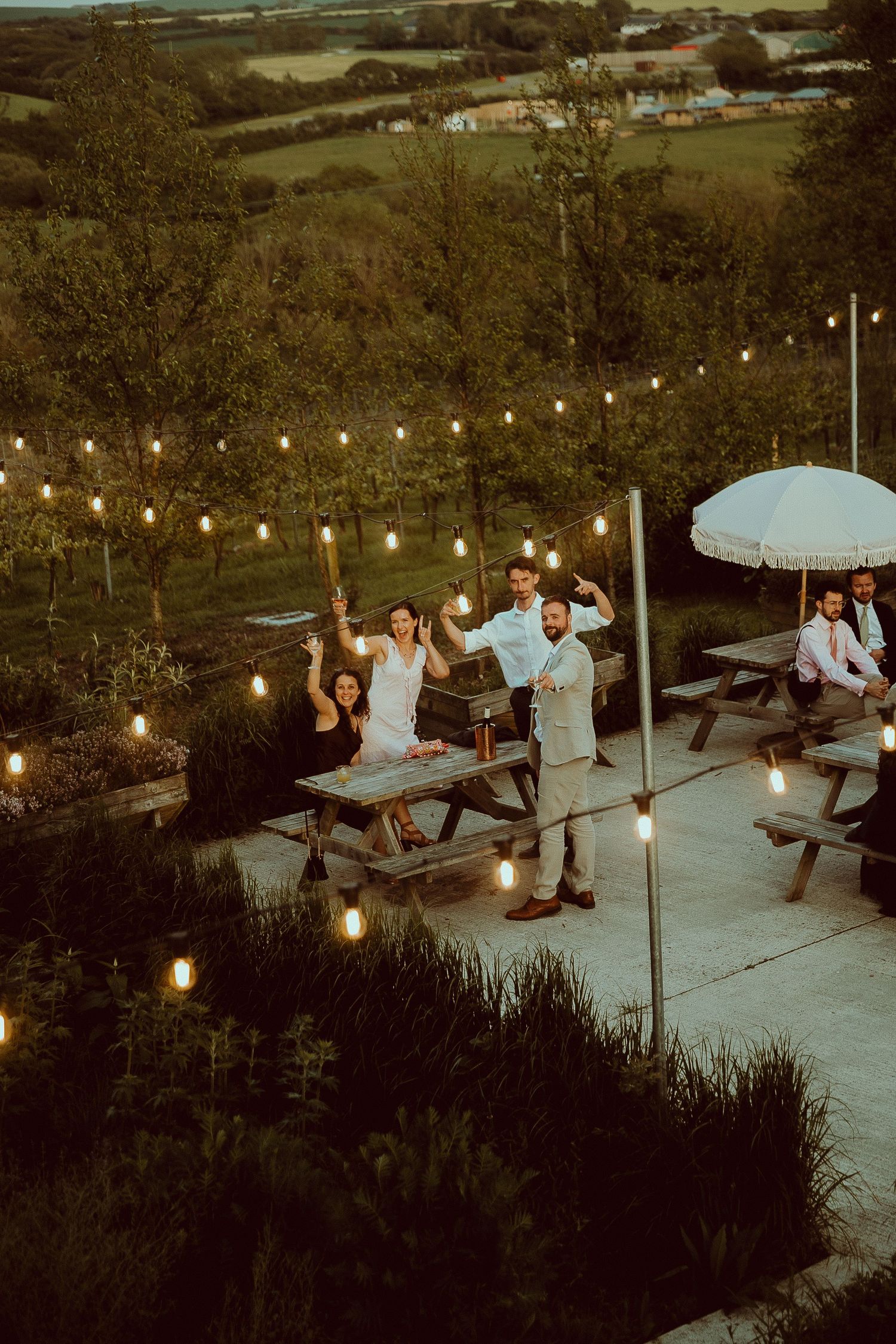 Aerial view of wedding guests celebrating under string lights at an outdoor venue with scenic countryside views at dusk.