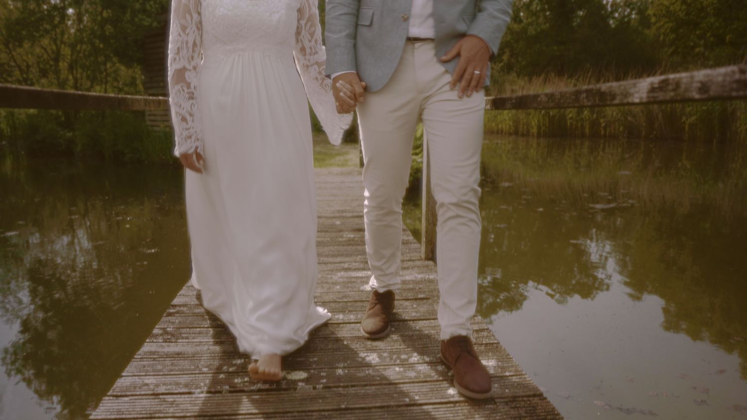 Couple in wedding attire holding hands on a wooden dock over a calm reflective pond surrounded by lush greenery.