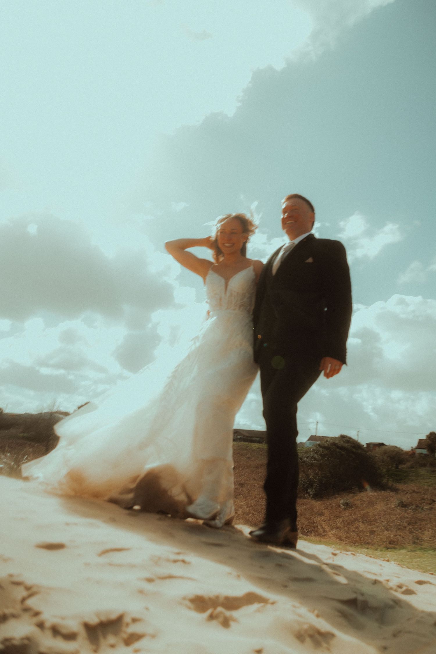 Bride in a flowing white lace wedding gown and groom in a dark suit pose together on a sandy dune beneath a bright, cloudy sky.