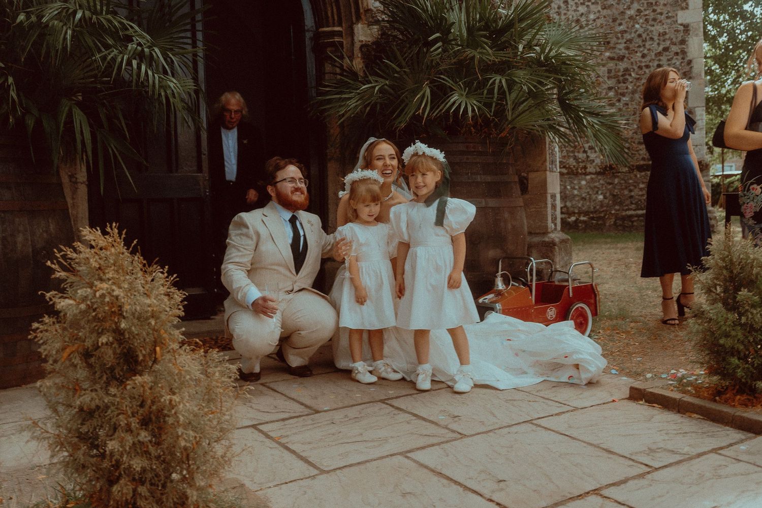 Groom crouching with flower girls in white dresses outside a rustic wedding venue surrounded by lush greenery.