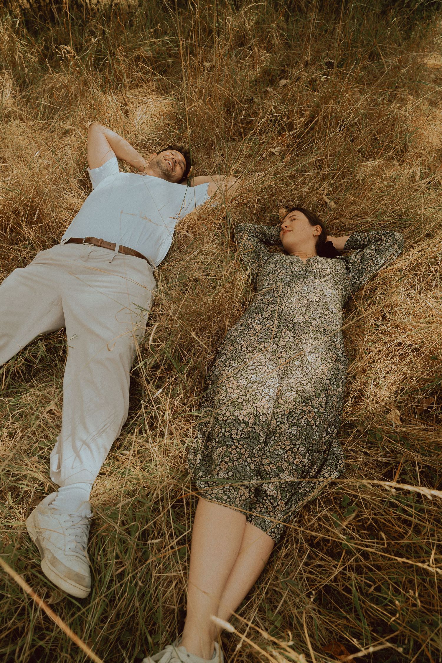 A couple lying in dry golden grass, relaxing outdoors in casual and floral attire during a serene countryside photo session.