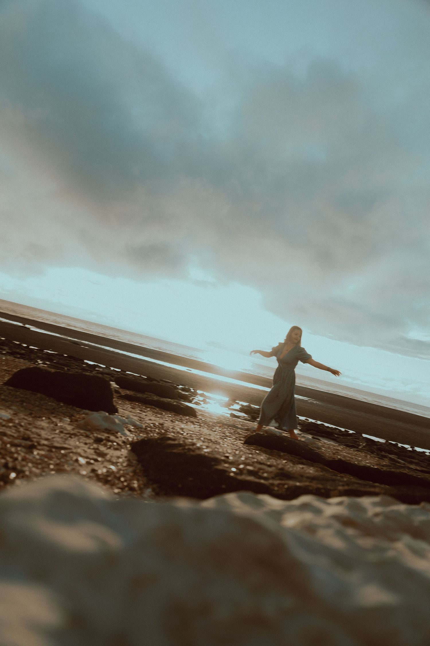Woman in a long dress stands with arms outstretched on a rocky beach at sunset under a dramatic cloudy sky.