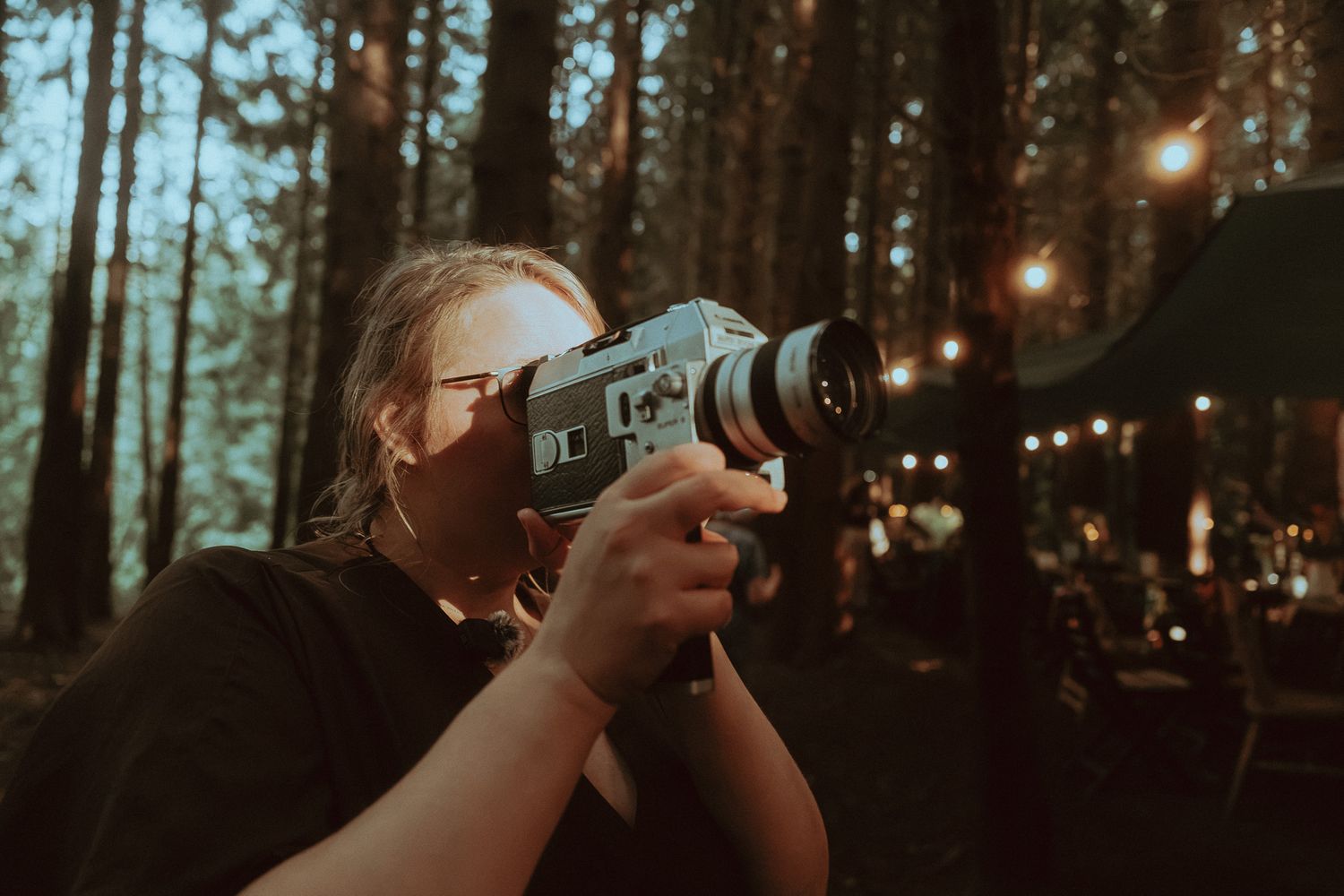 Person filming with a vintage Super 8 camera in a forest setting with bokeh lights in background.
