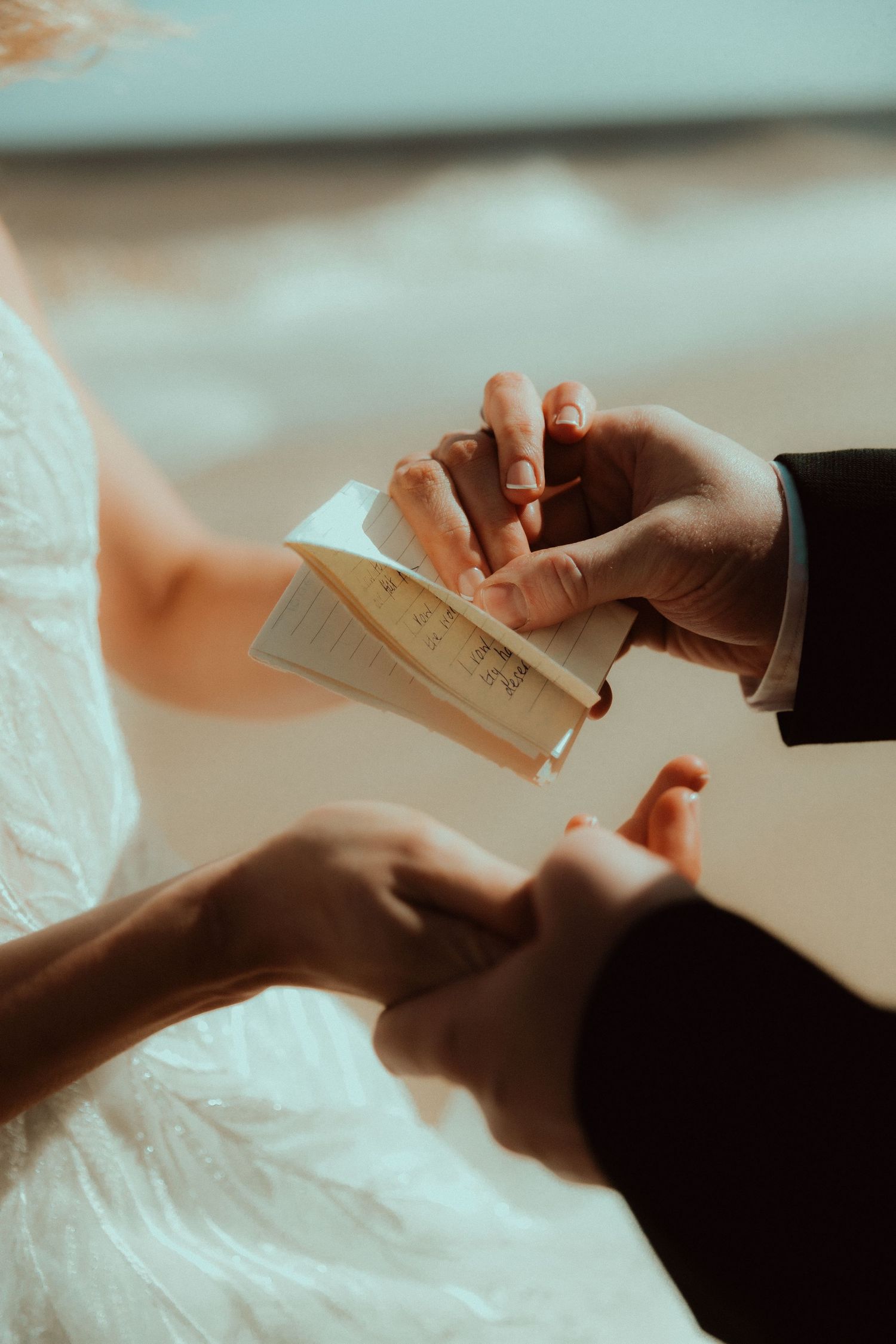 Couple exchanging wedding vows outdoors, groom in suit holding vow booklet while bride in white dress holds hands.