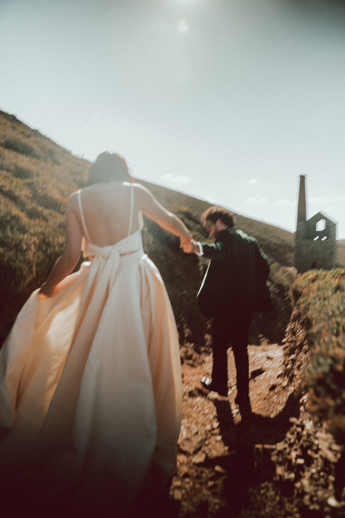 Bride and groom holding hands walking up a sunlit hillside path, bride in elegant white gown, rustic building in background.
