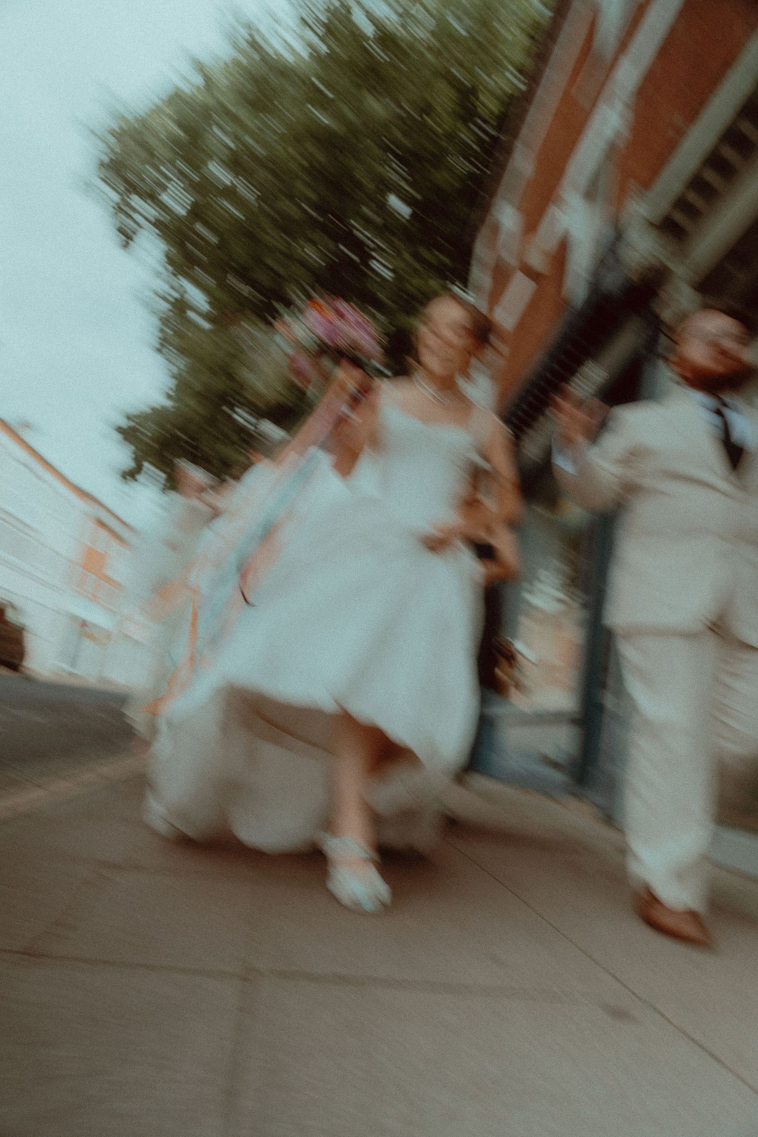 Blurred motion photo of a bride in white dress walking with wedding party on a sidewalk near brick building.