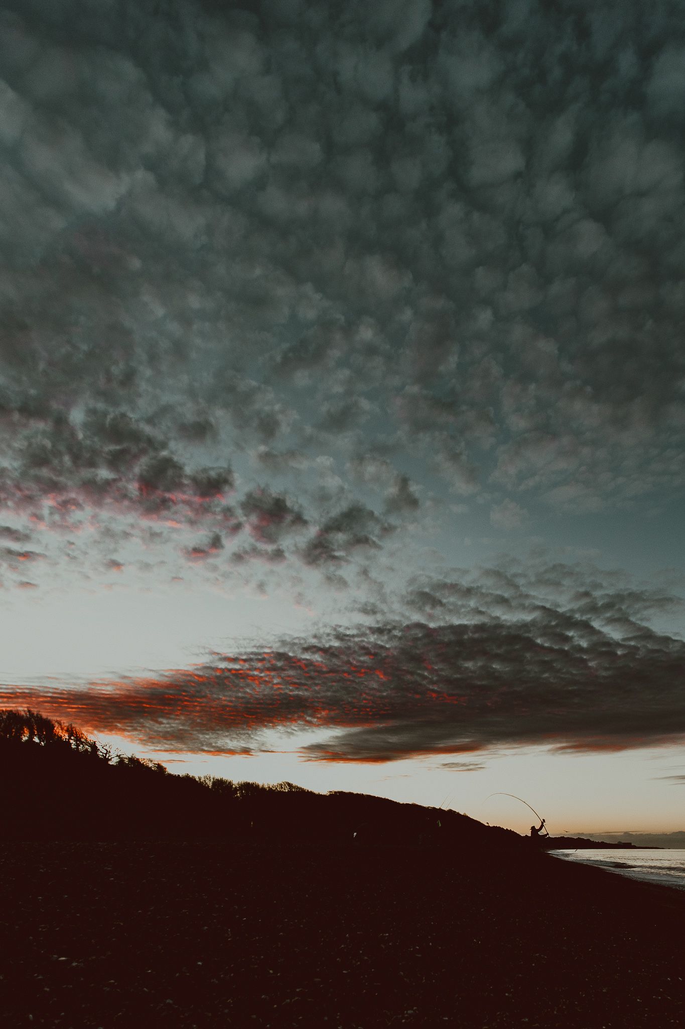 A fisherman silhouetted sea fishing during a dramatic sunset sky with textured altocumulus clouds glowing pink.