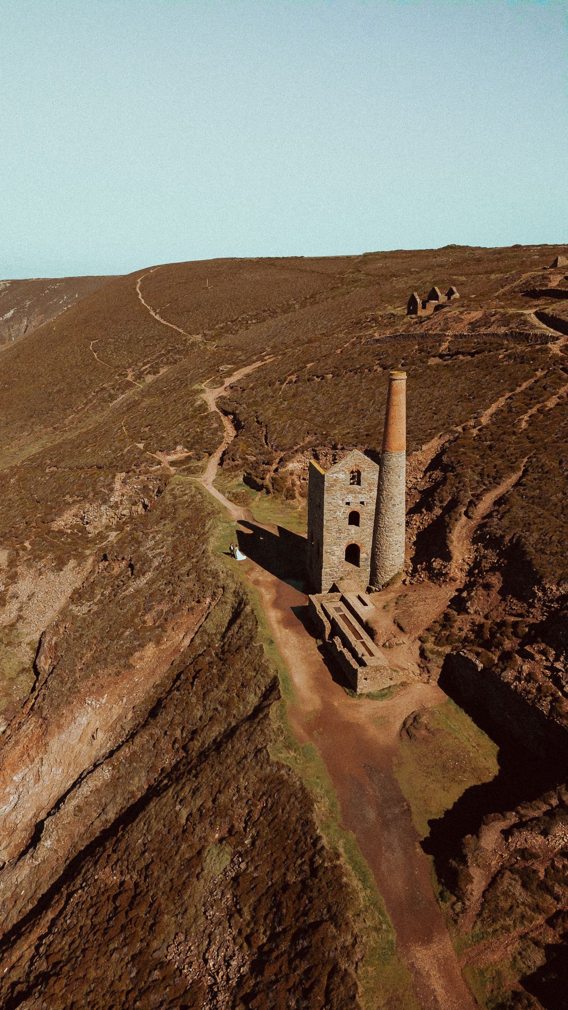 Aerial view of an old stone mining engine house perched on rugged cliffs along a winding dirt path in Cornwall.