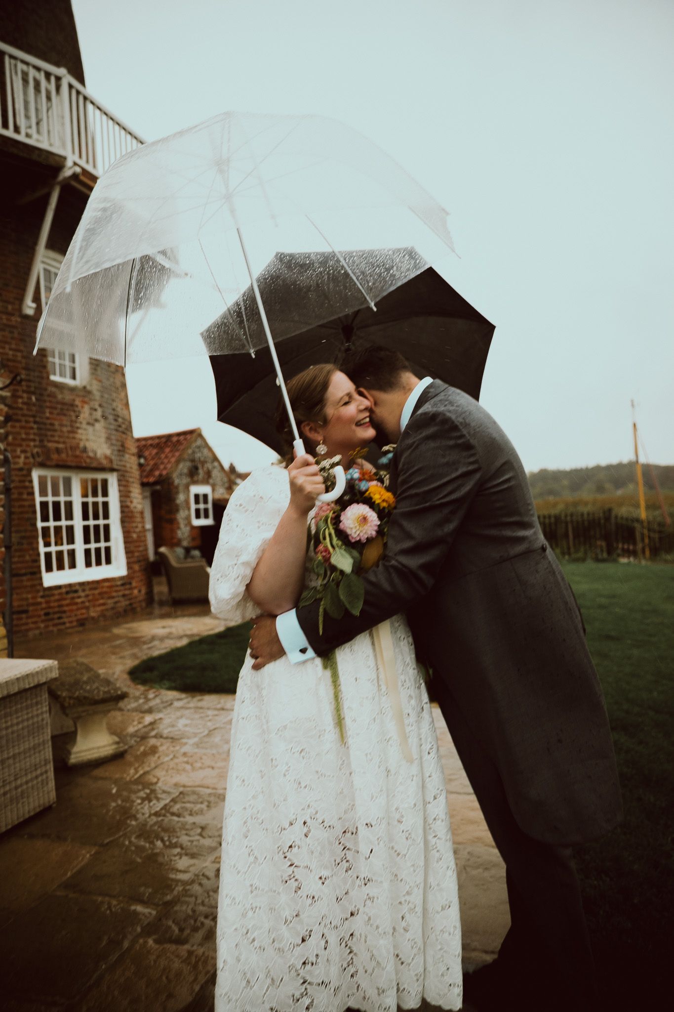 A romantic wedding moment under umbrellas on a rainy day outside Cley Windmill wedding venue at the North Norfolk coastal marshes.