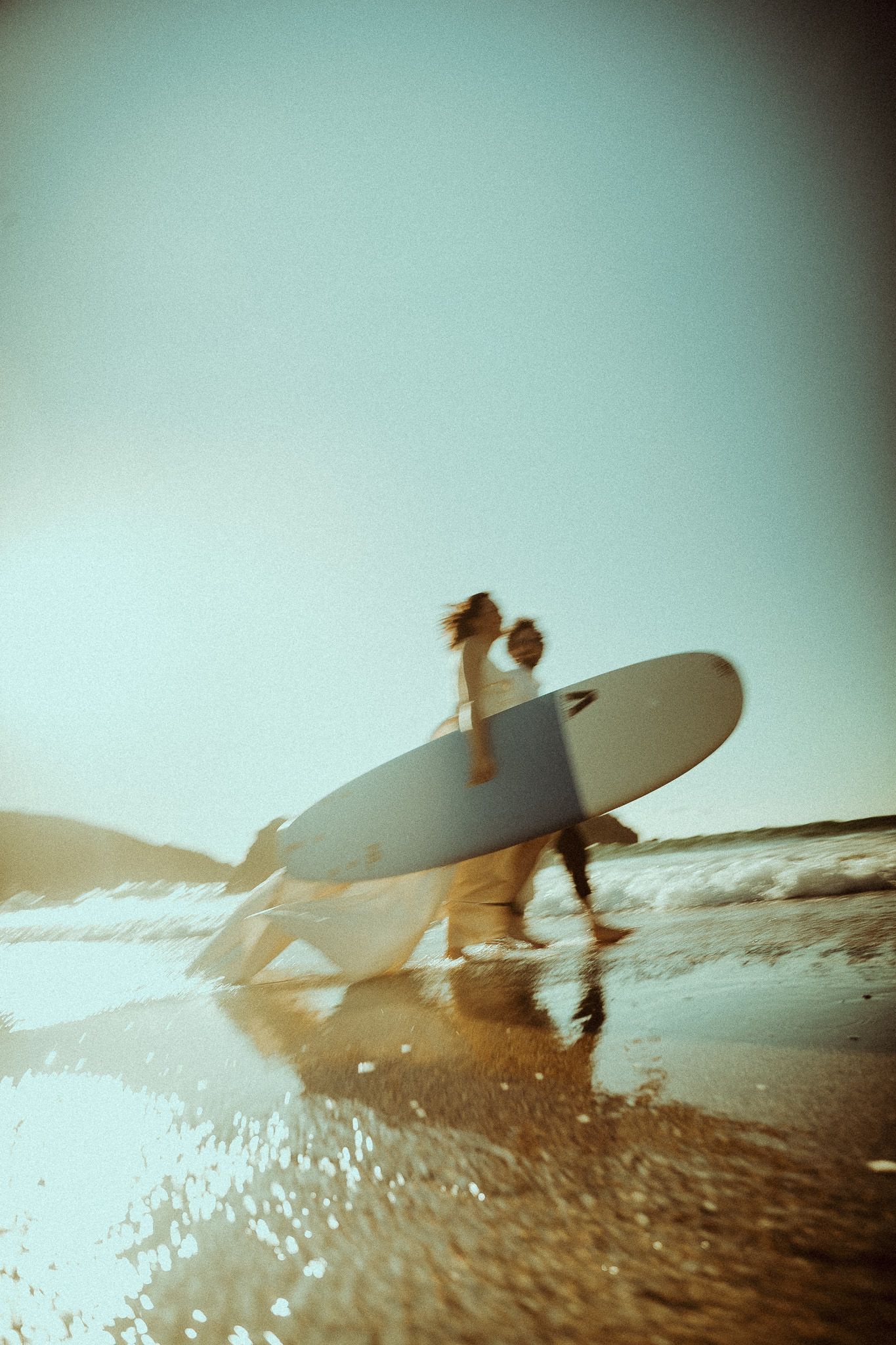 A newly married couple carrying a white surfboard along a sunlit beach shoreline, captured in a dreamy motion-blurred film photo.