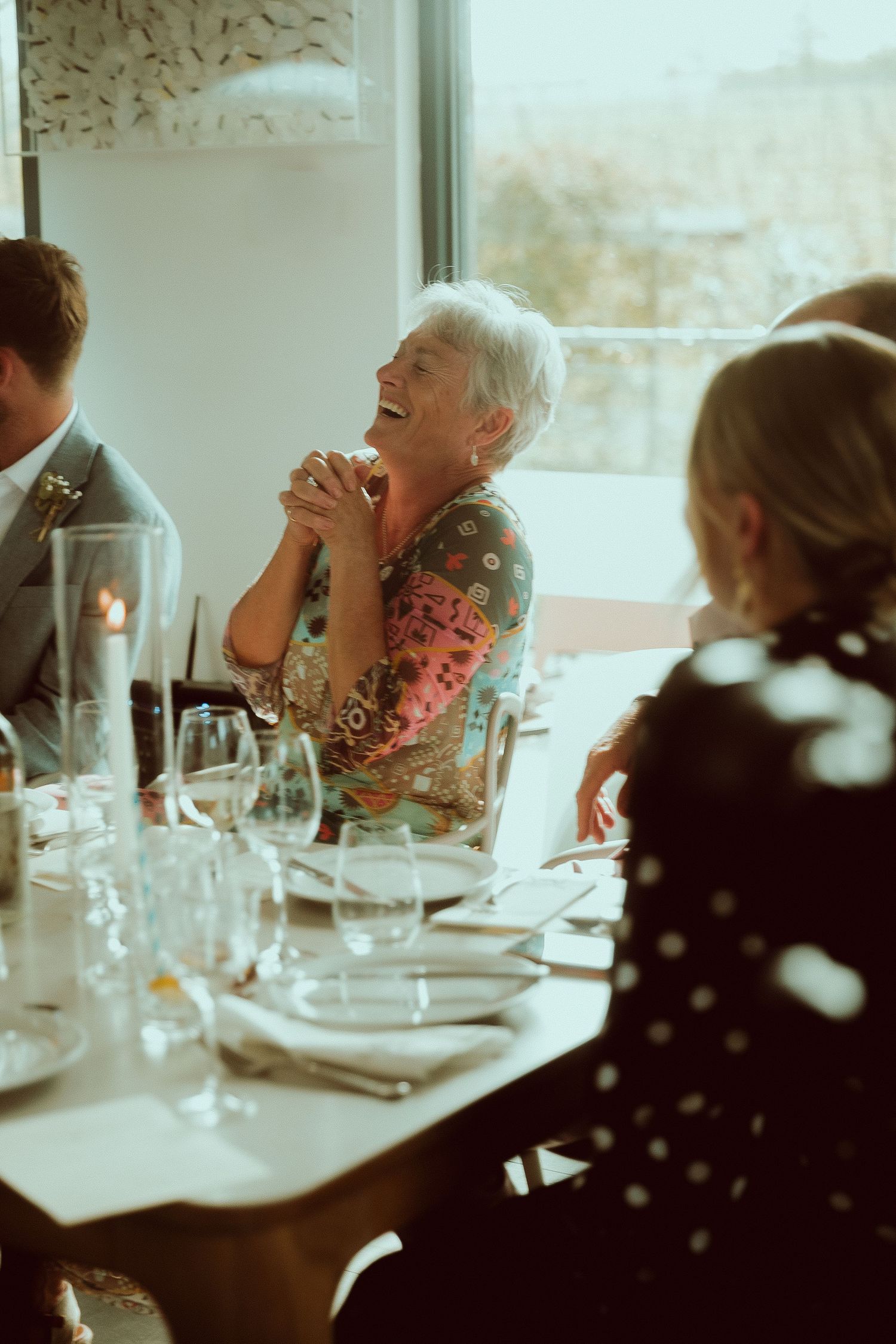 Elderly woman laughing joyfully at a formal dinner table, wearing a floral blouse, surrounded by guests.