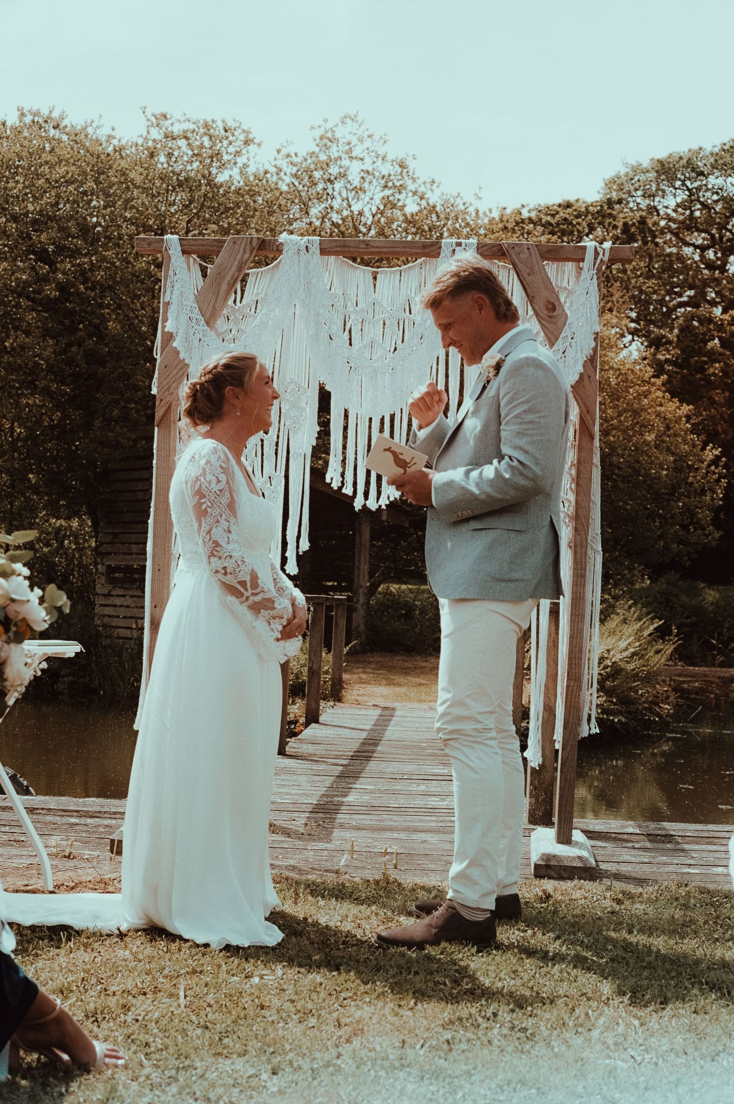 Couple exchanging vows outdoors under a macrame arch near a pond, bride in lace gown, groom in light gray suit.
