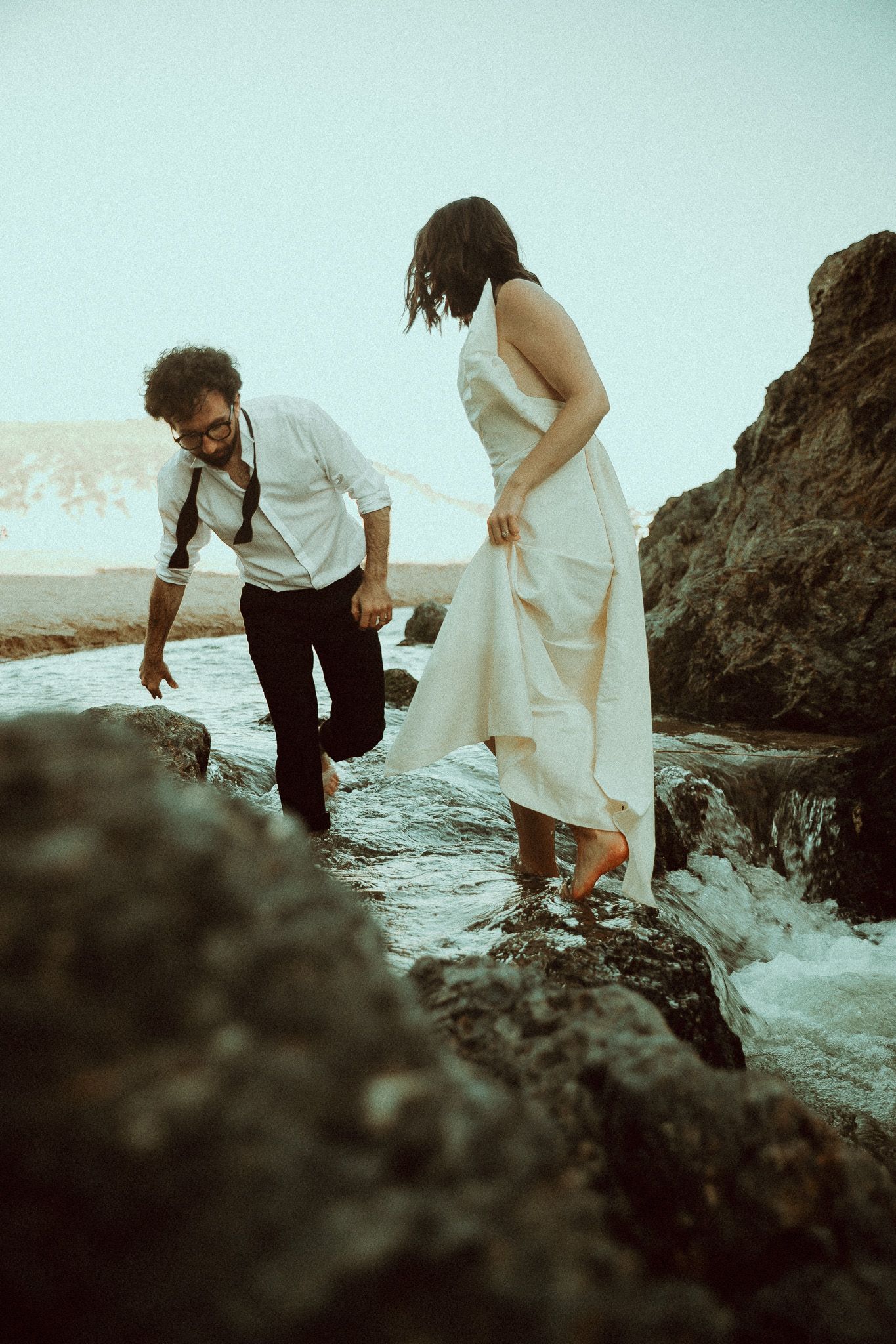 Barefoot couple in a white dress and rolled-up trousers wade through shallow ocean water between dark coastal rocks at dusk.