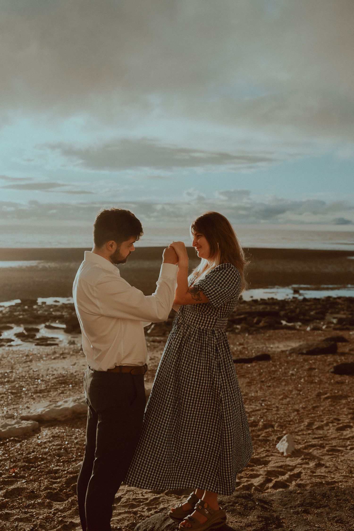 Romantic couple holding hands at sunset on a rocky beach, man in white shirt and woman in plaid dress.