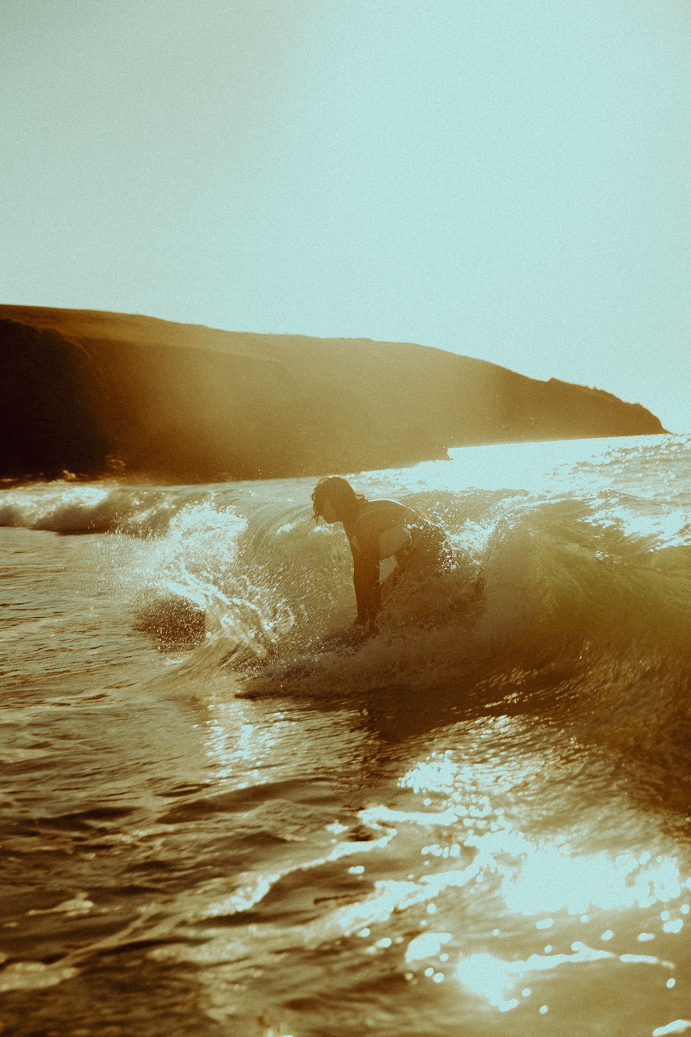 Surfer leaning into a breaking wave along a rugged coastline, bathed in warm golden sunset light and sparkling ocean spray.
