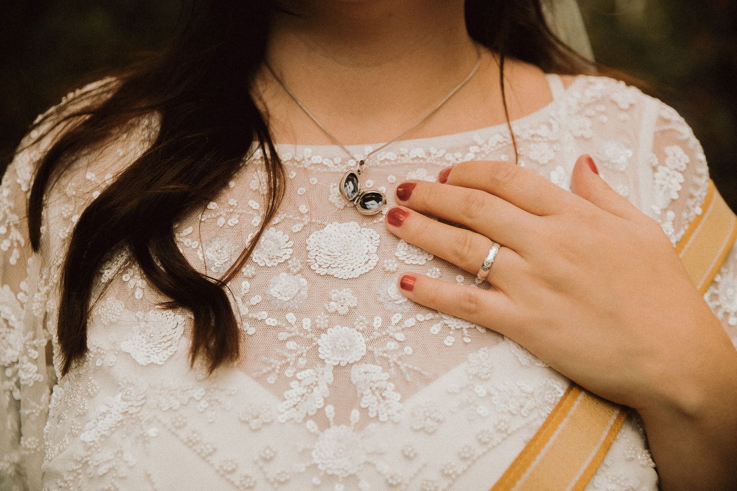 Woman in white lace dress with red nails, silver ring, and delicate necklace, hand resting on chest.