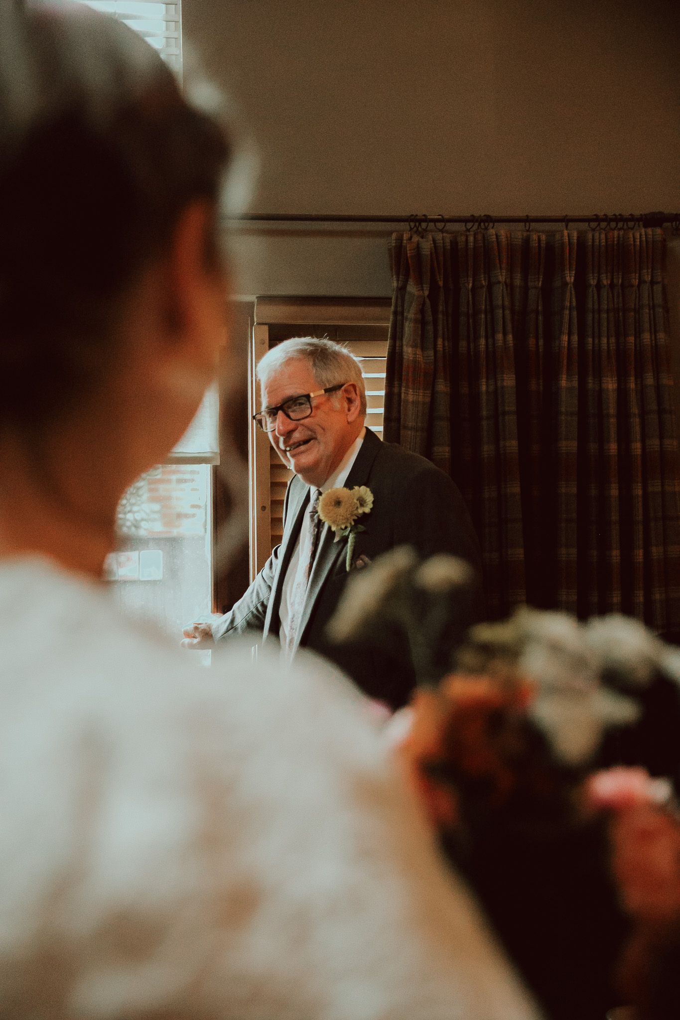 Emotional father seeing bride in wedding dress for first time, wearing boutonniere, glasses, smiling warmly.