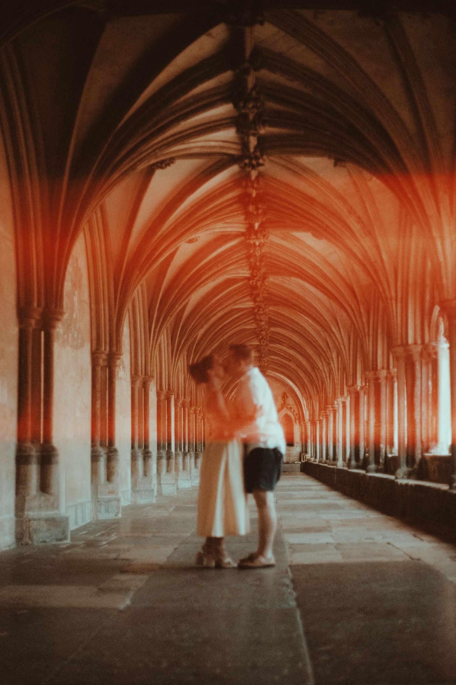 A couple shares a romantic kiss in a Gothic cathedral cloister with stunning ribbed vaulted ceilings bathed in warm red tones.