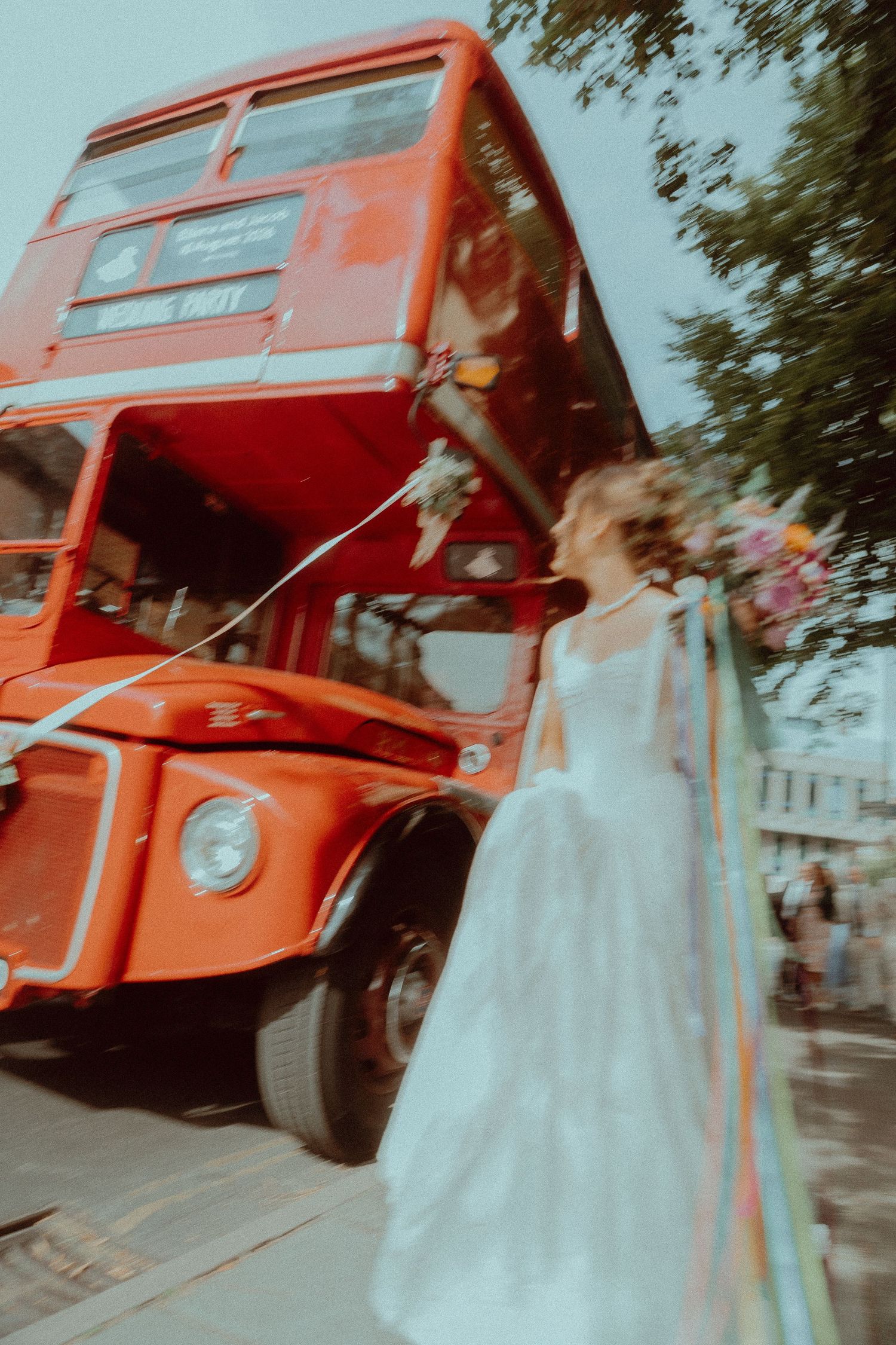 Bride in white gown holding colorful bouquet beside a classic red London double-decker bus on a city street.