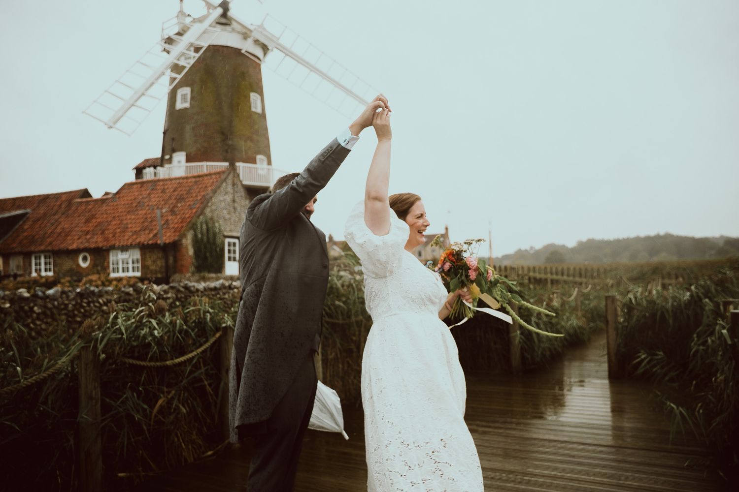 Couple dancing joyfully in front of a historic windmill on a rainy wedding day in the countryside.