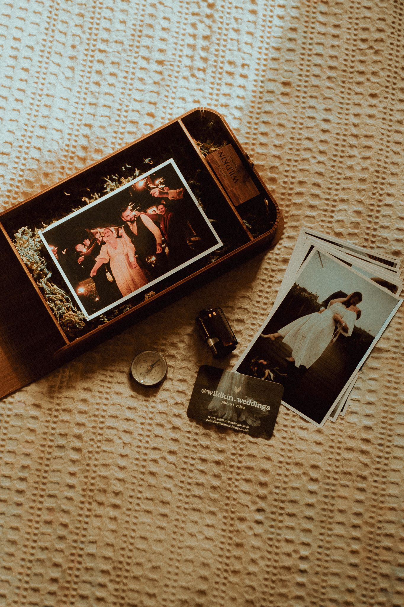 Rustic wooden box with wedding photos, film roll, compass, and photographer business cards on white knit blanket.
