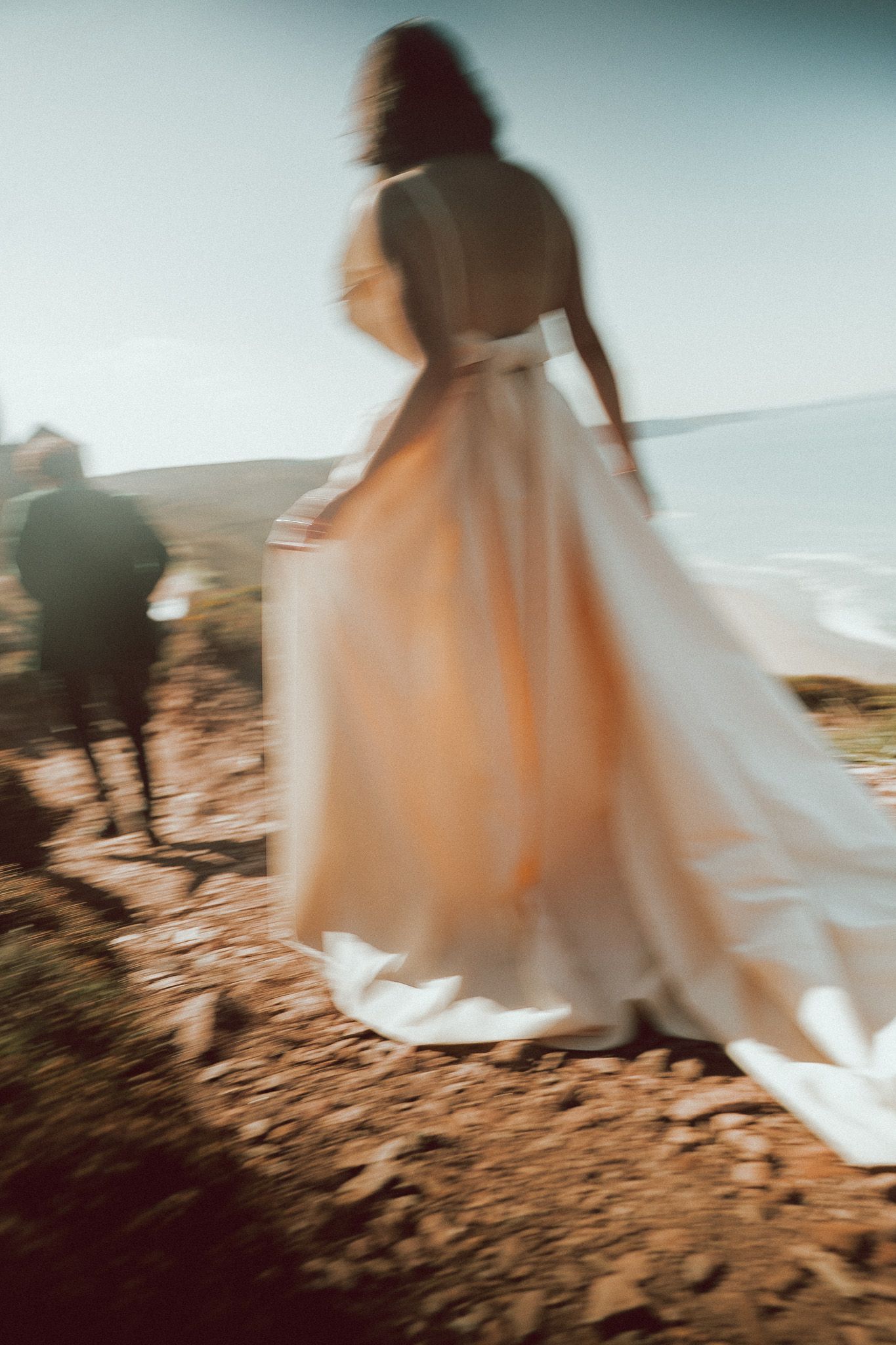 Blurred motion shot of a bride in a flowing backless gown walking along a coastal cliff with a horse nearby.