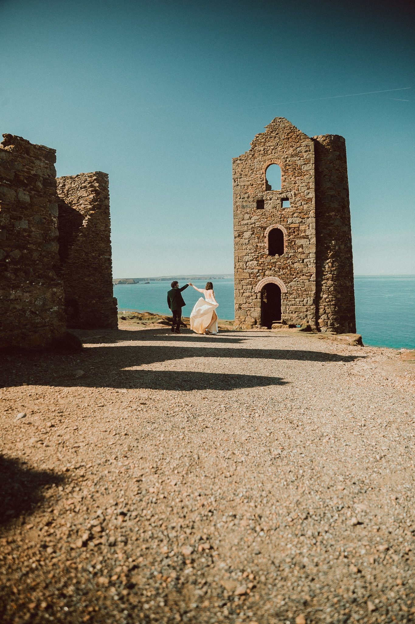 Bride in a flowing white gown dances with her partner near ruined stone engine houses on a sunny clifftop overlooking the blue sea.