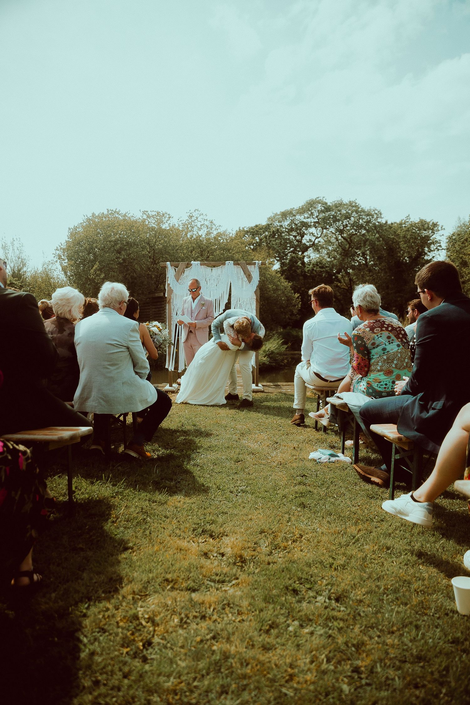 Outdoor wedding ceremony on grass aisle with guests seated, couple kissing at altar with officiant and floral backdrop.