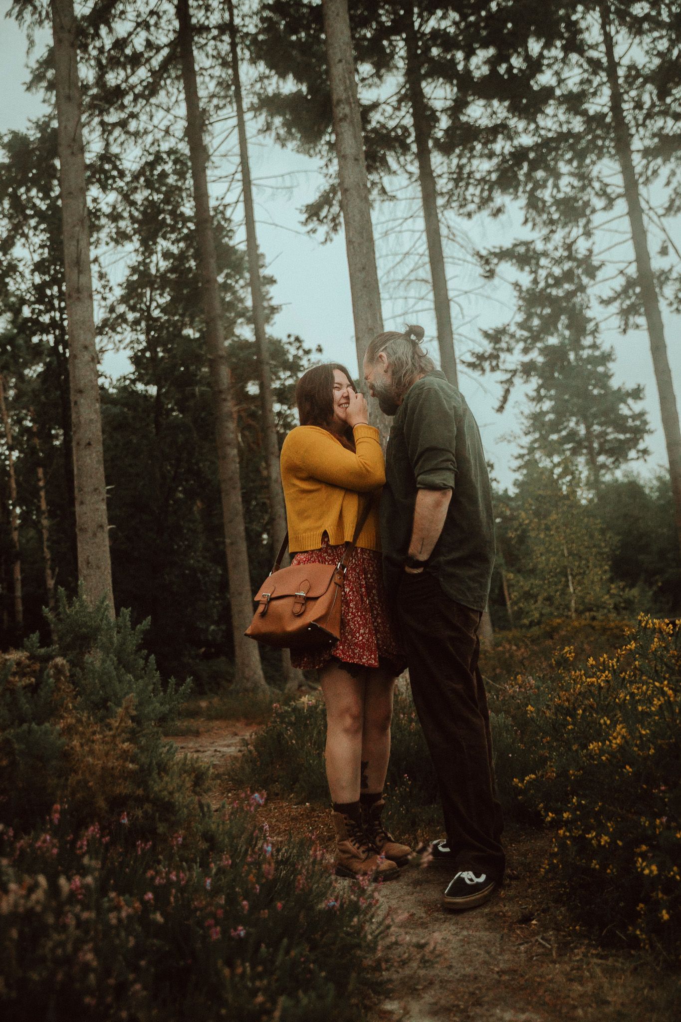 Two people share a romantic moment while hiking through a misty forest trail surrounded by wildflowers and tall trees.