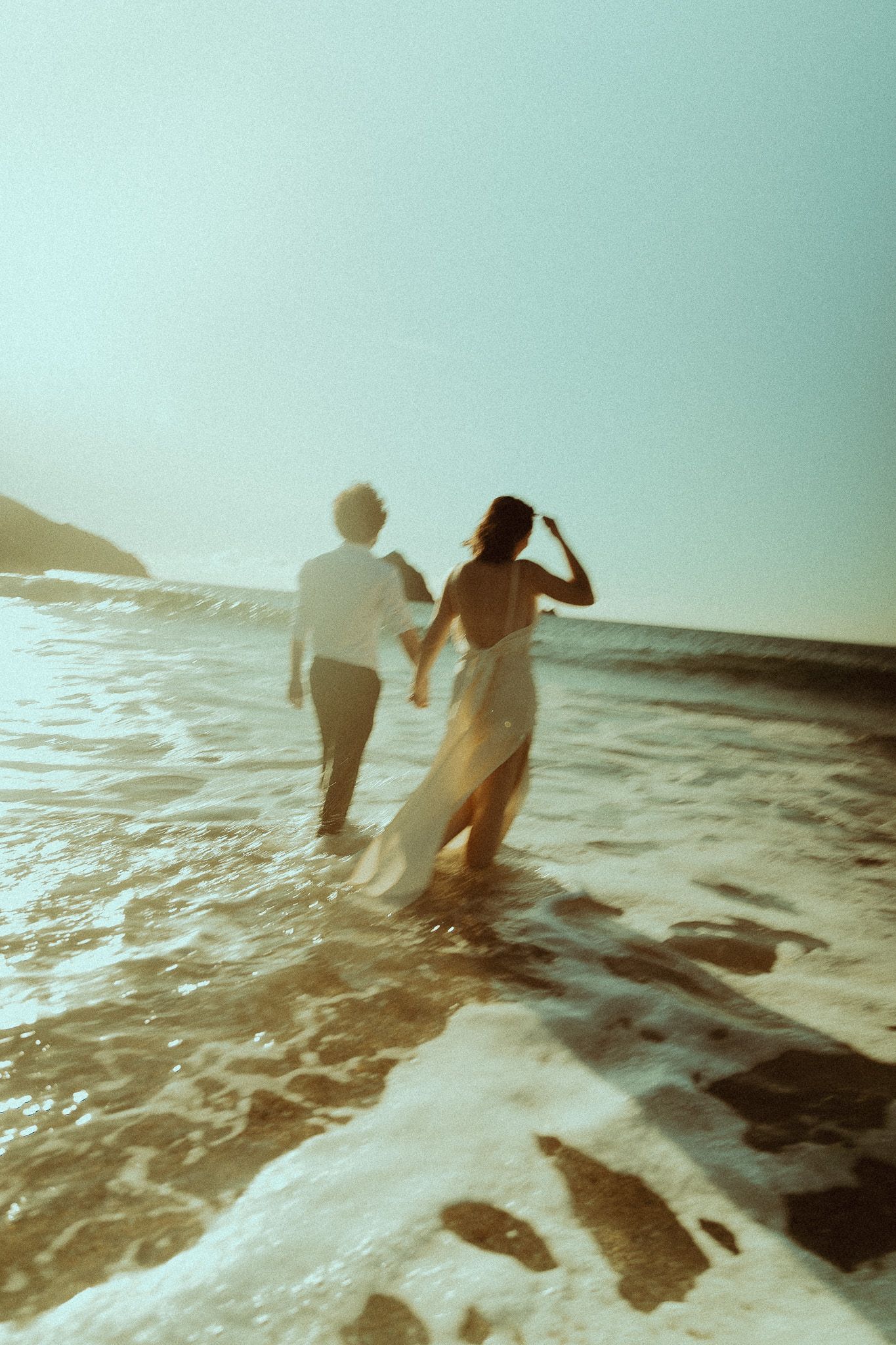 Couple holding hands as they wade through shallow ocean waves at sunset, in a dreamy, sun-drenched beach photo with soft motion blur.